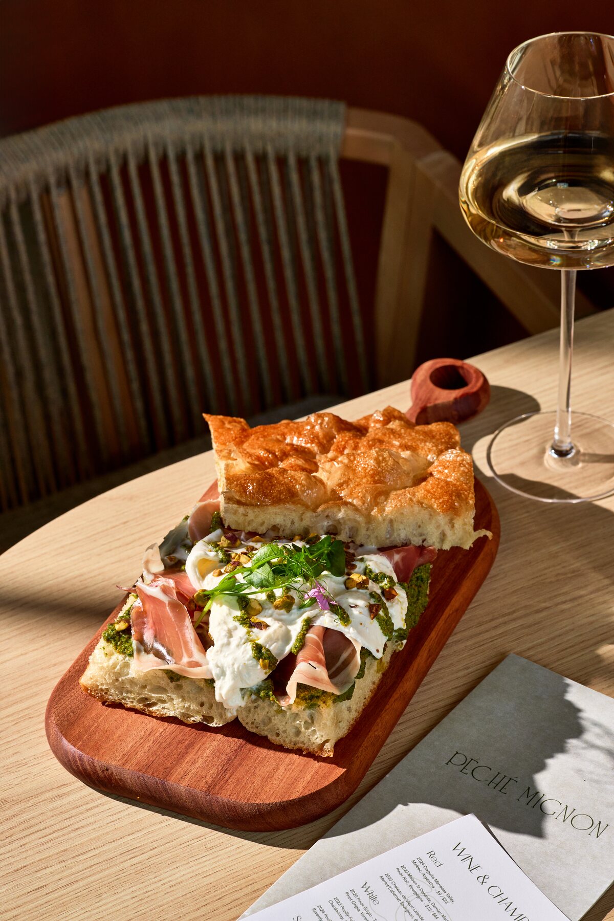 Italian sandwich with prosciutto, burrata, pistachio pesto, and arugula on focaccia beside a glass of white wine at Péché Mignon